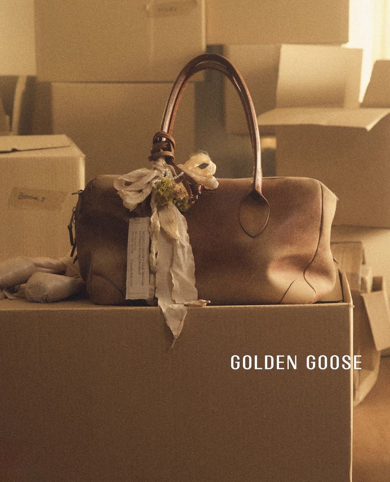 Golden Goose brown distressed leather bag with ribbon embellishments resting on cardboard moving boxes.