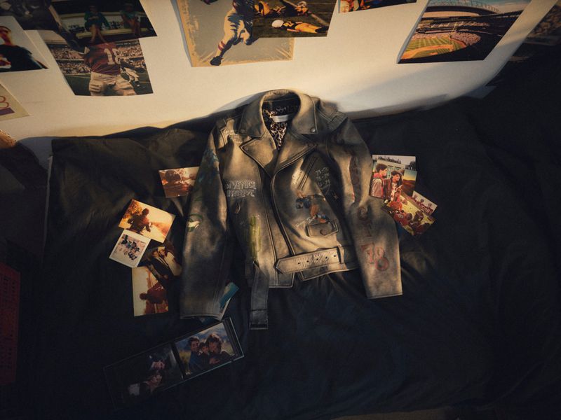 Distressed grey leather biker jacket with custom markings laid flat on dark bedding alongside scattered photographic prints.