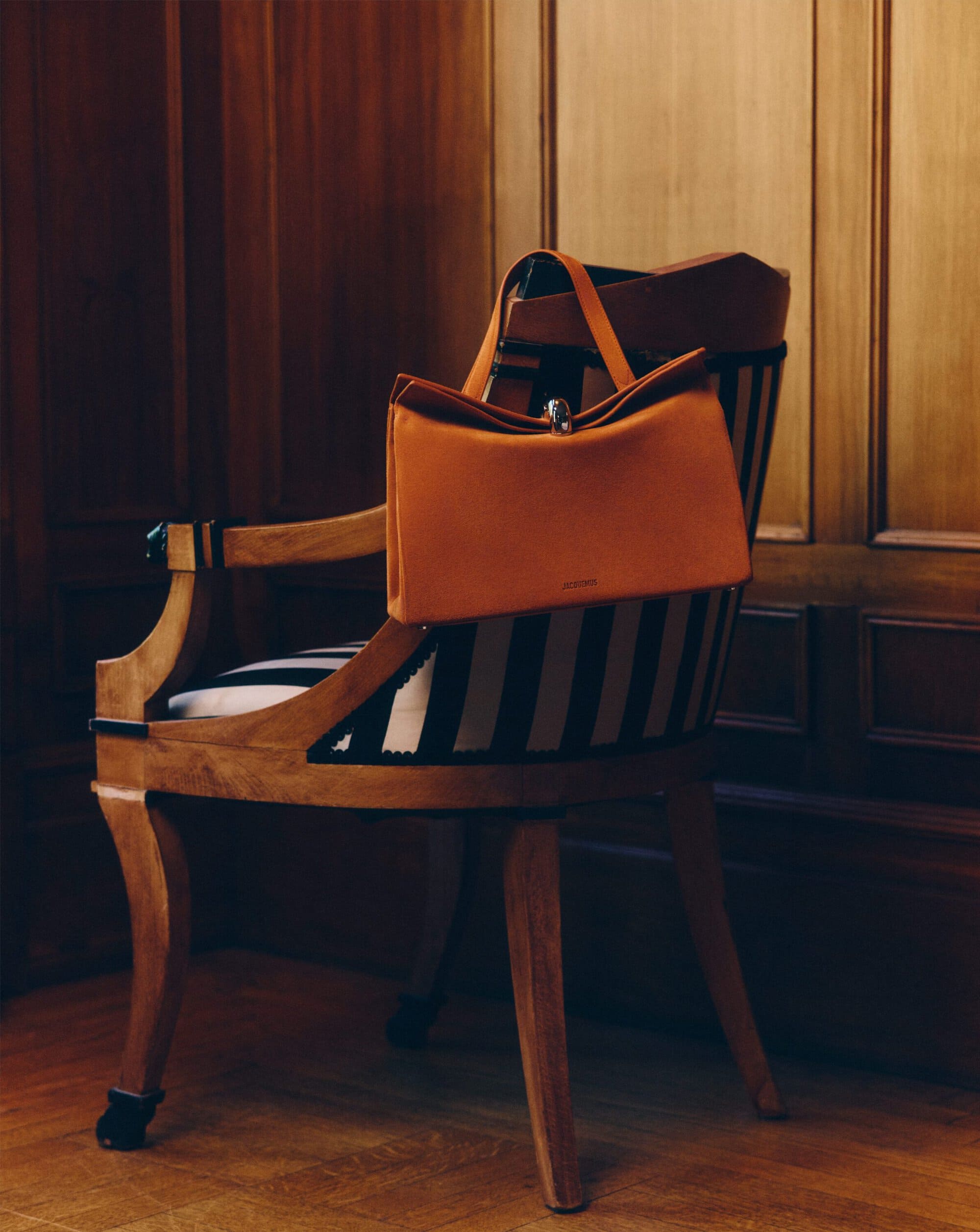 Orange structured leather handbag draped over the back of a black-and-white striped wooden armchair in a wood-paneled room.
