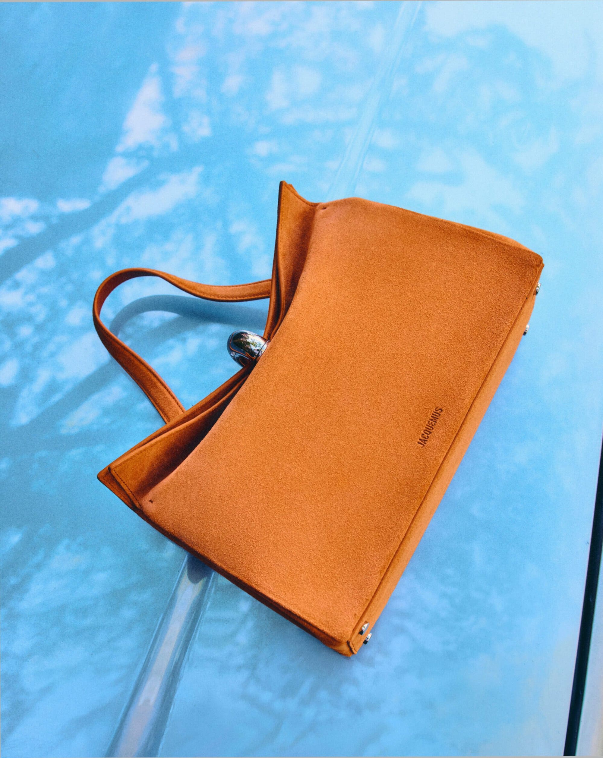 Jacquemus orange suede handbag resting diagonally on a reflective light blue surface with outdoor tree shadows.