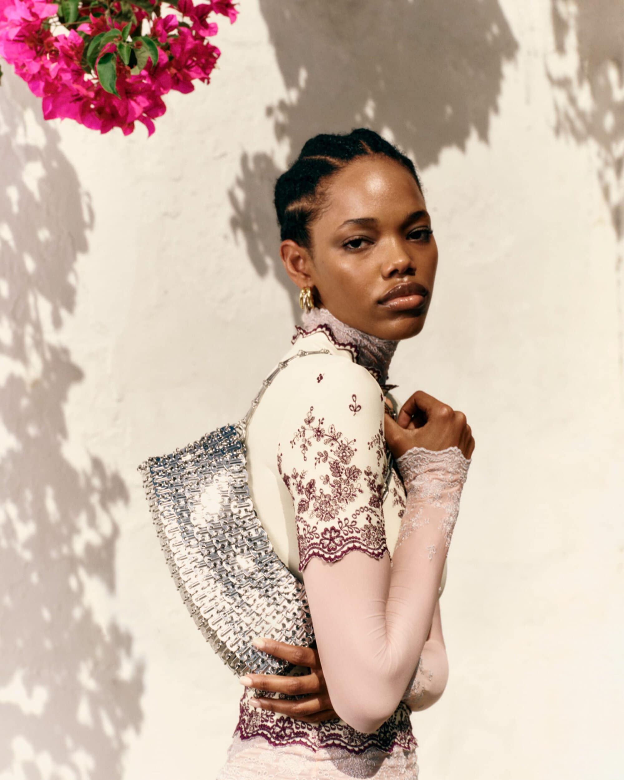 Black woman in a lace-trimmed high-neck top holding a silver metallic mesh shoulder bag against a sunlit wall with pink blossoms.