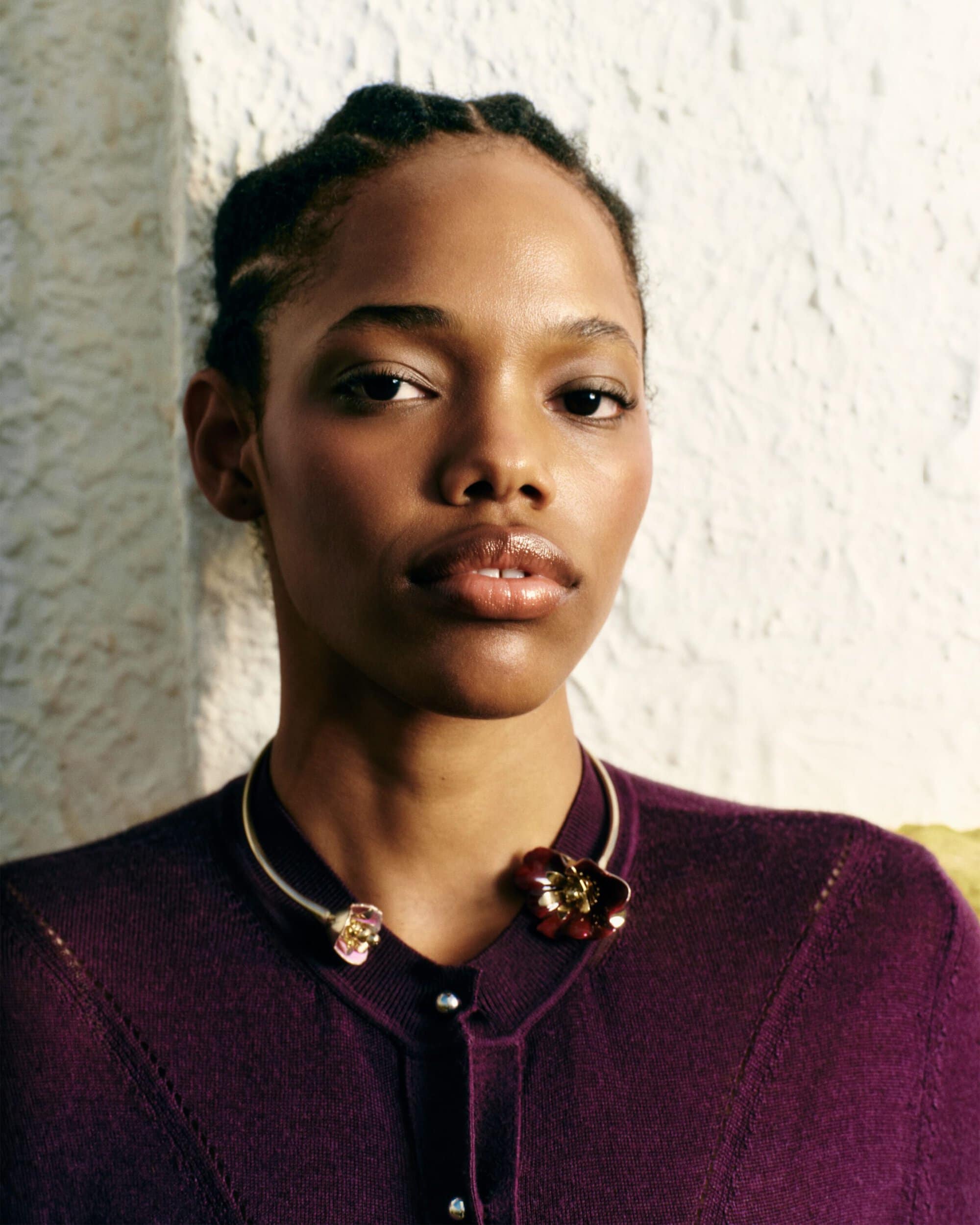 Portrait of an African American woman wearing a maroon cardigan and floral statement necklace against a textured wall backdrop.
