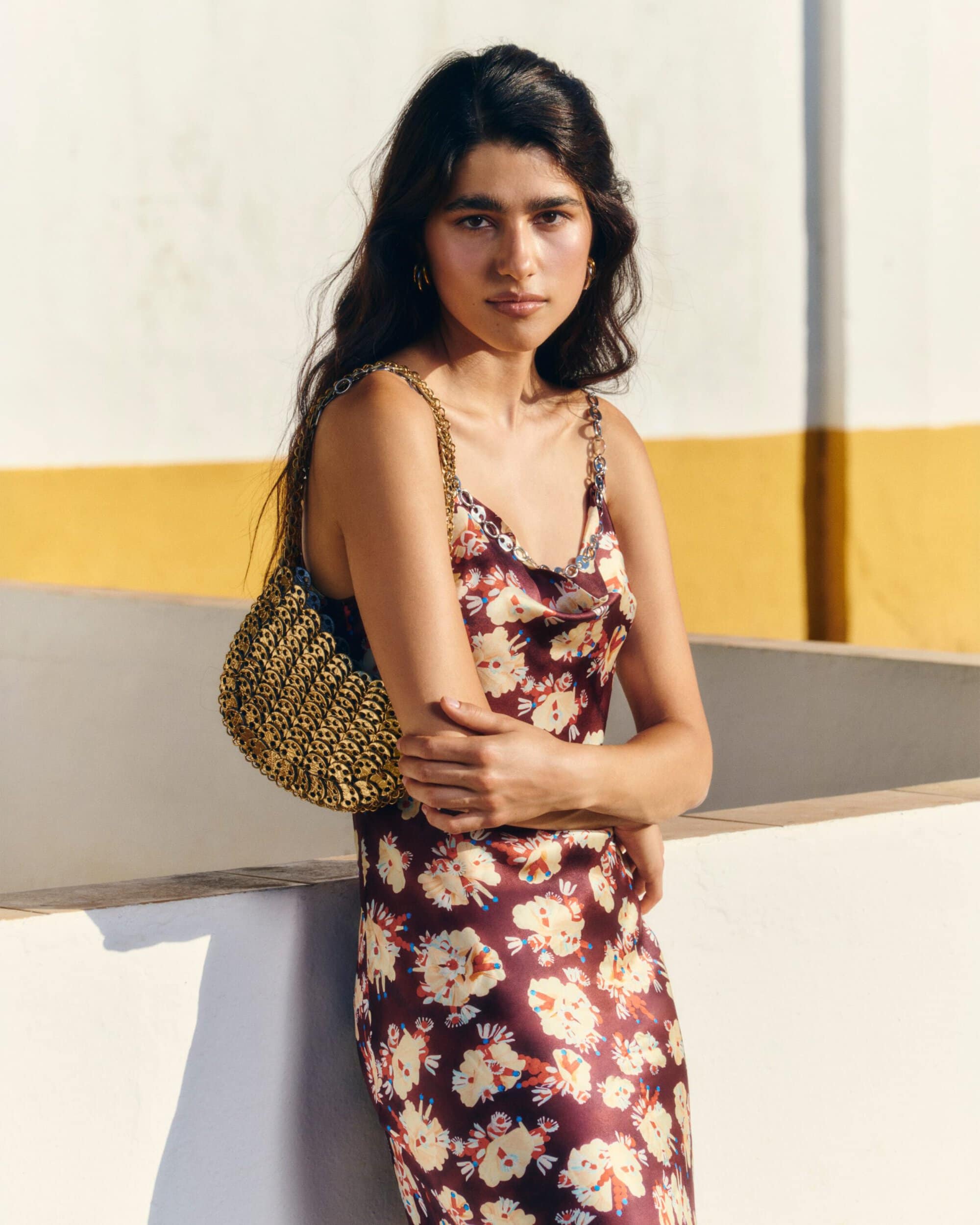 Model wearing a floral satin slip dress and gold chainmail shoulder bag against a white and yellow wall in direct sunlight.
