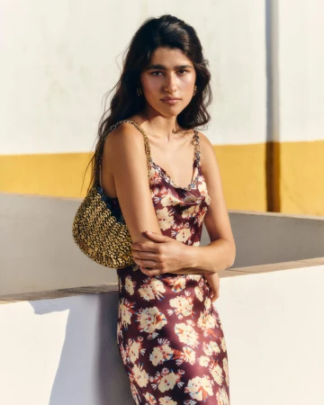 Model wearing a floral satin slip dress and gold chainmail shoulder bag against a white and yellow wall in direct sunlight.