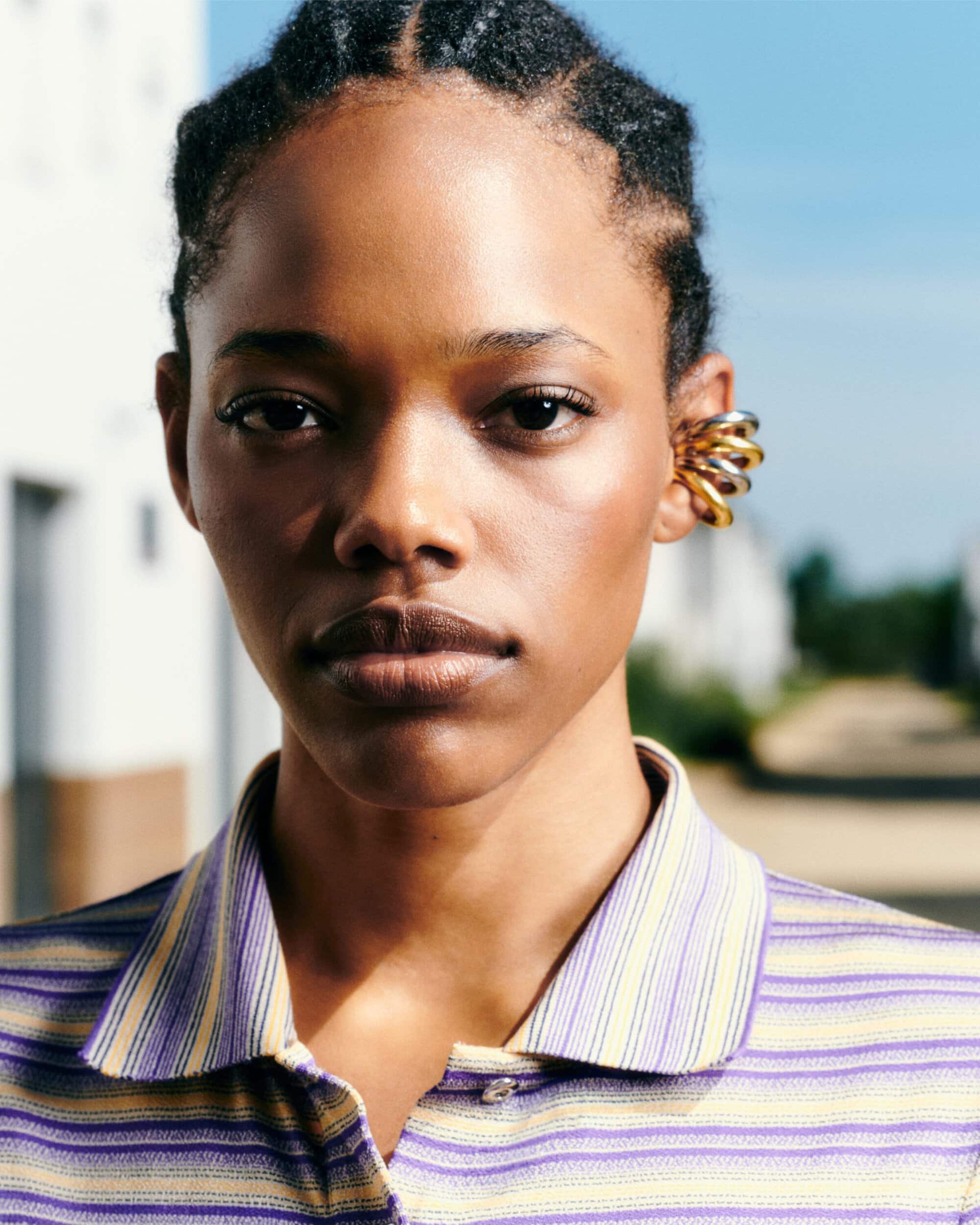 Close-up portrait of a Black woman with braided hair wearing a striped lavender polo shirt and stacked gold and silver ear cuff.