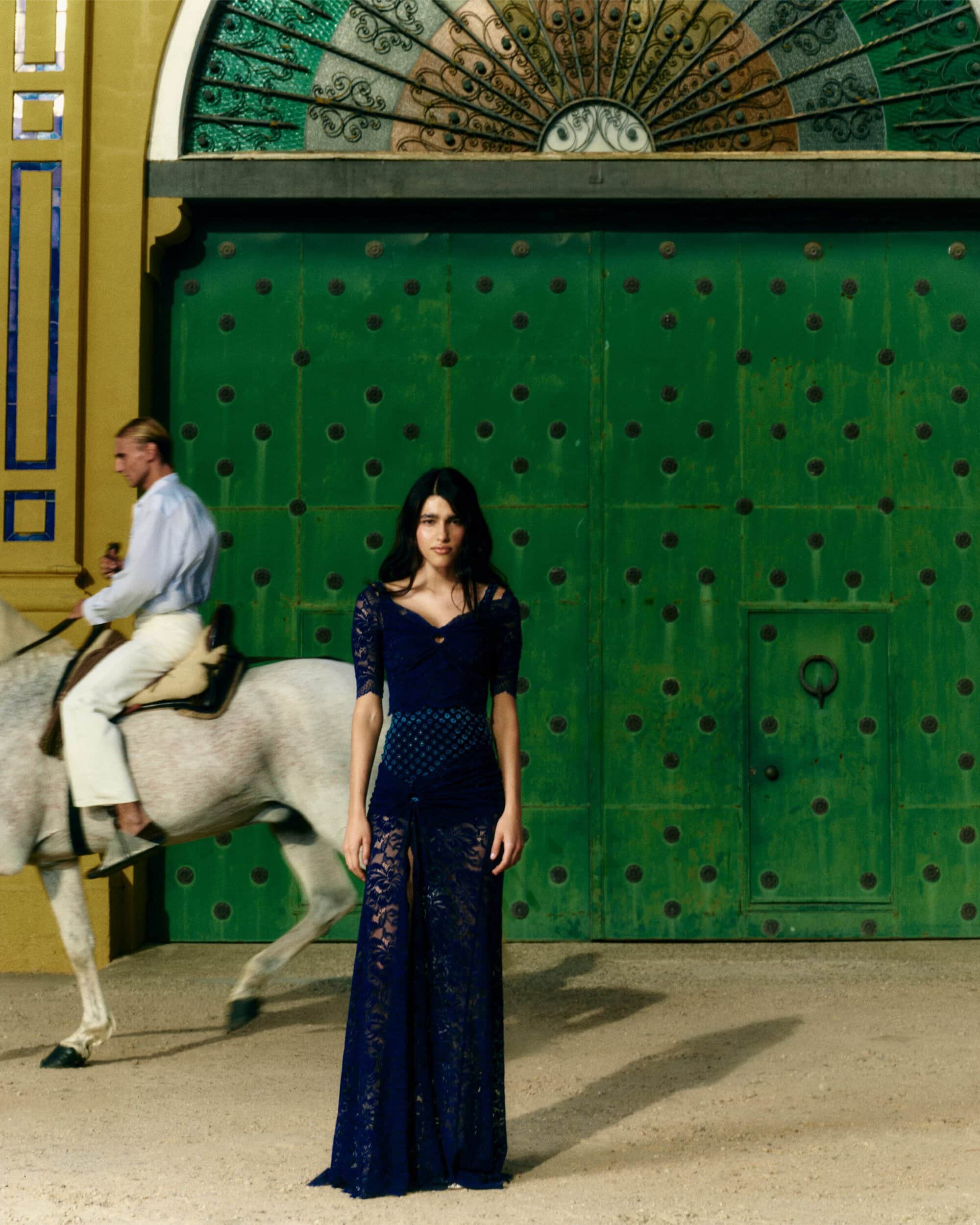 Woman in a sheer navy lace gown standing before large green arched doors with a man on a white horse behind her.