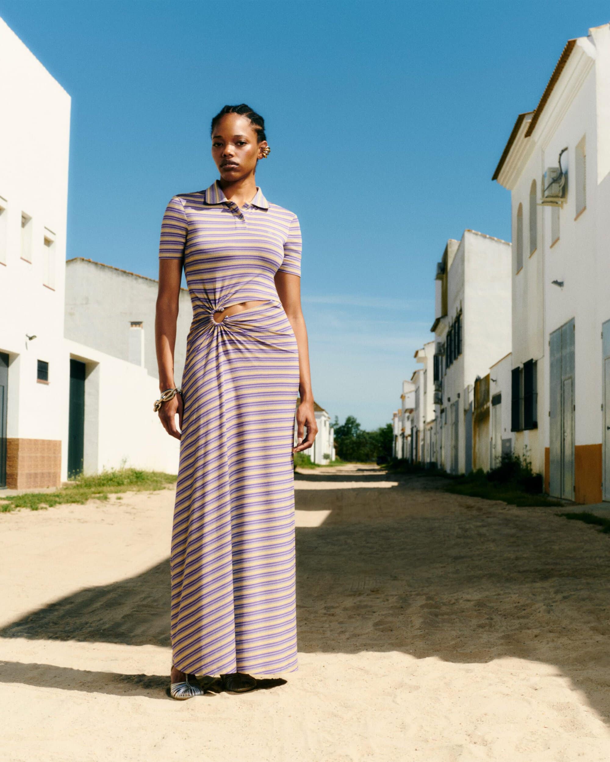 Model standing outdoors on a sandy village street wearing a striped maxi polo dress with a waist cut-out ring detail.
