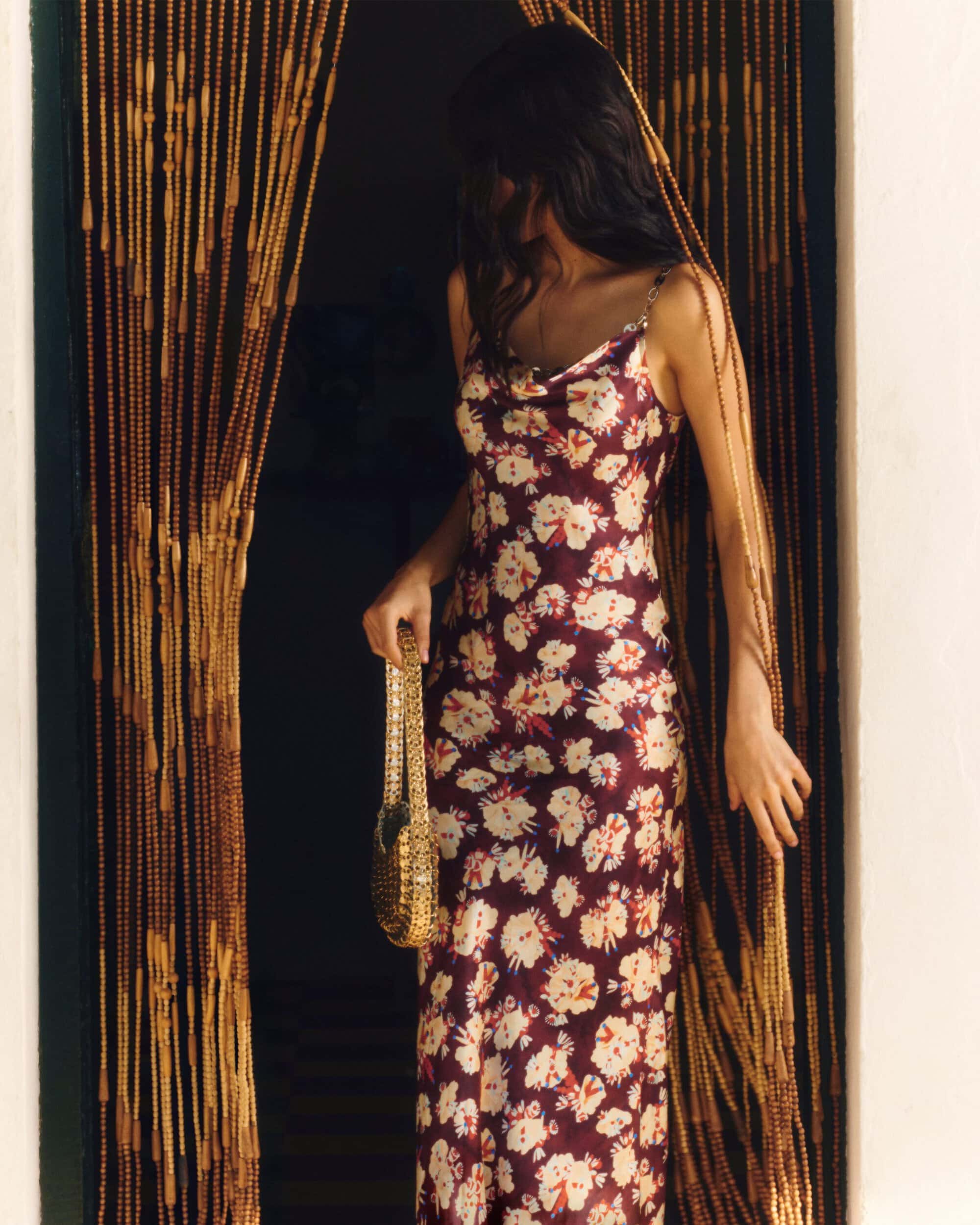 Woman in a floral burgundy slip dress holding a gold metallic handbag standing between hanging beaded curtains.