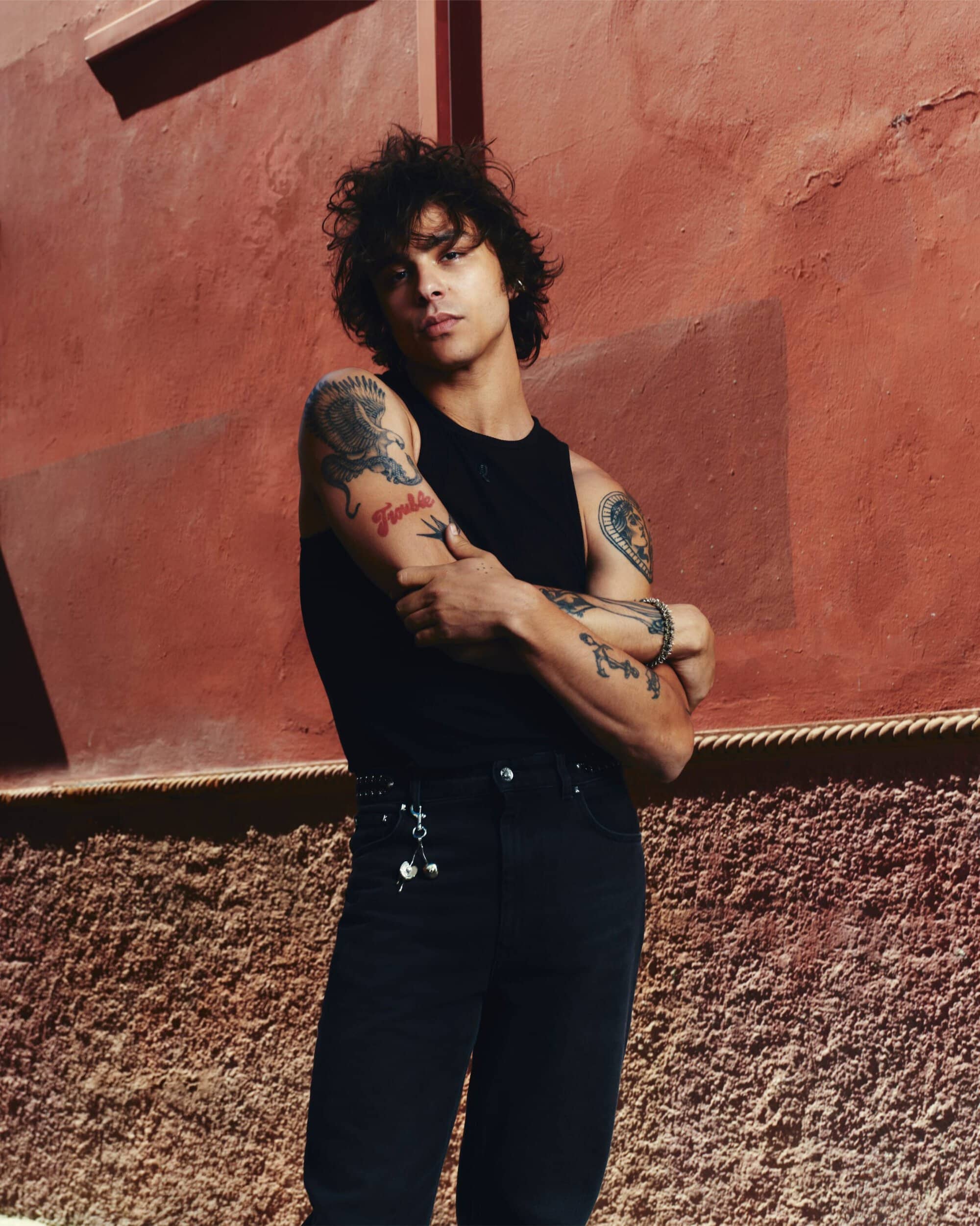 Man with tattoos wearing a black sleeveless tank top and black pants standing outdoors against a textured terracotta wall.
