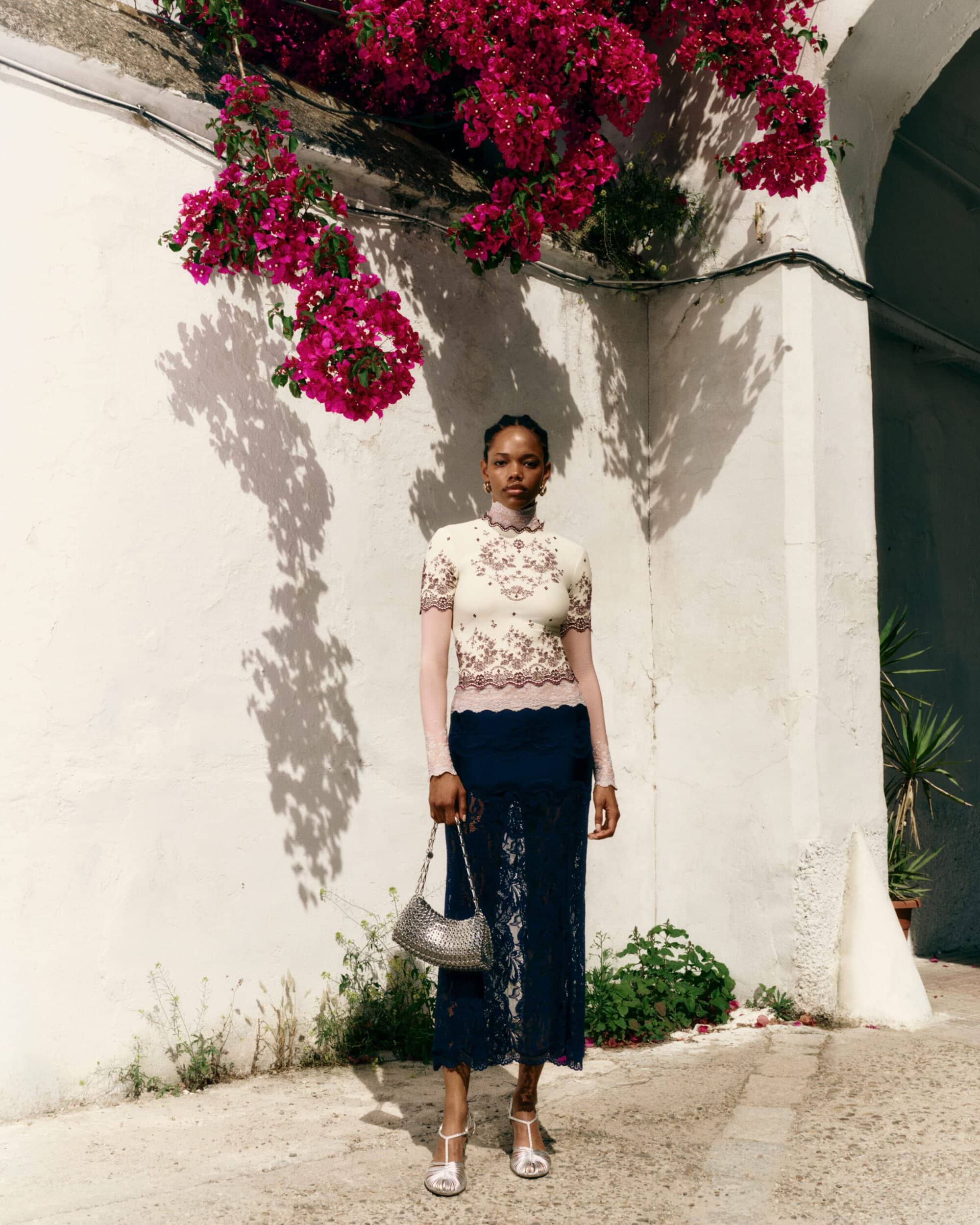 A woman in an embroidered cream top and navy lace skirt holding a silver chainmail bag under magenta bougainvillea by a white wall.