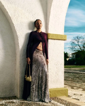 Model posing under a white stucco archway wearing a deep plum open cardigan, shimmering silver maxi skirt, and gold chain bag.