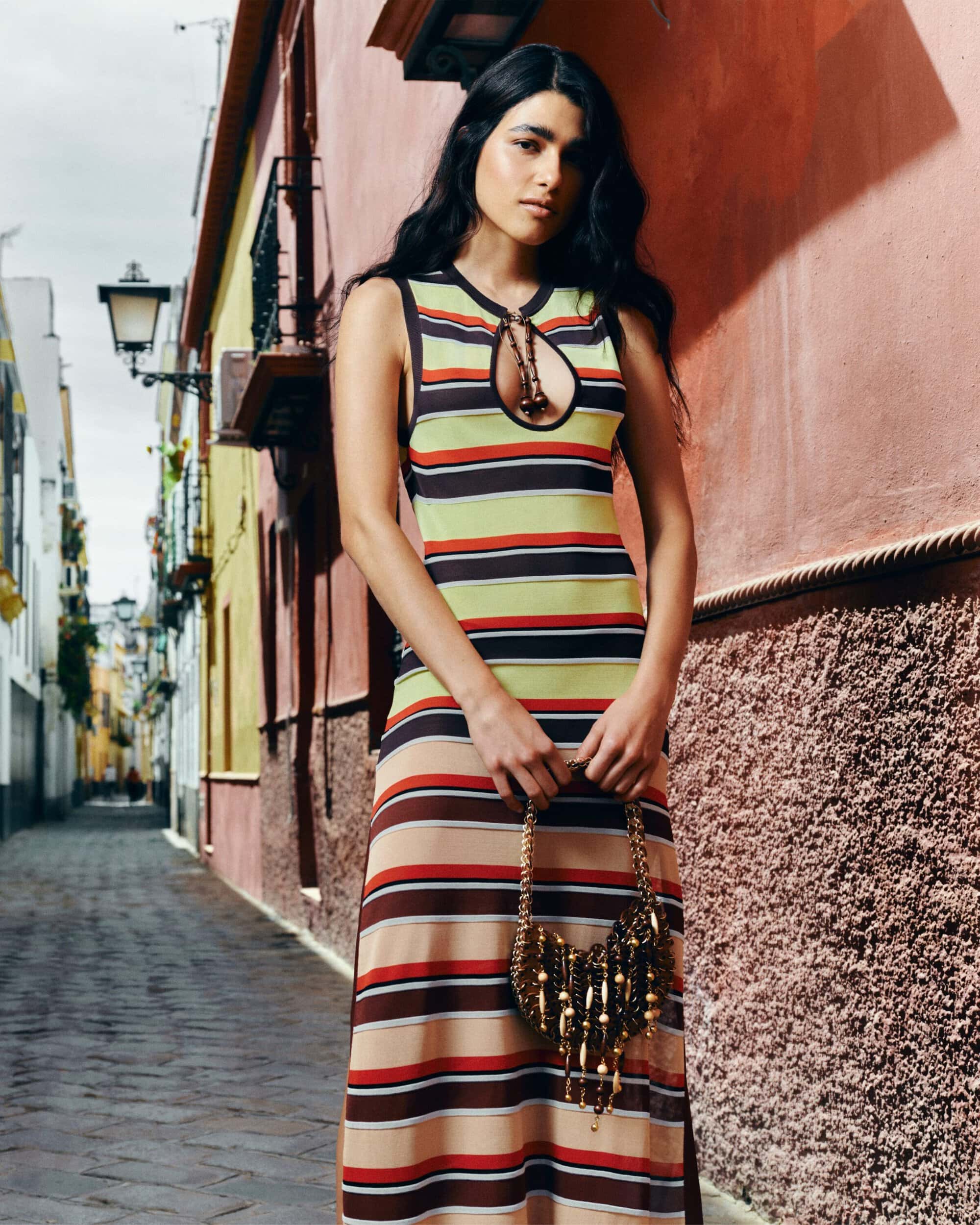 Model in a striped sleeveless dress holding a beaded handbag standing in a narrow urban street