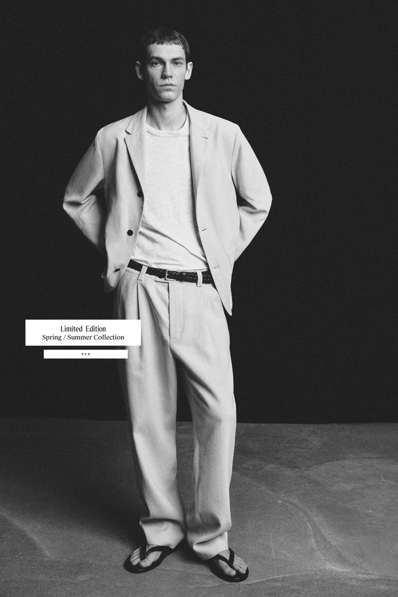 Aleksandr Gordeev poses in a light suit jacket, white t-shirt, loose trousers, and sandals against a dark studio background.