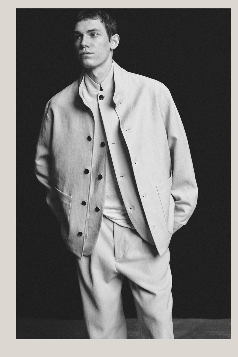 Portrait of an adult male wearing a light-colored jacket and pants against a dark background in a studio setting; fashion shot.