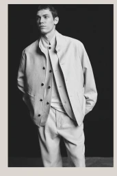 Model Aleksandr Gordeev poses in a layered light-colored jacket and matching trousers against a black studio backdrop.