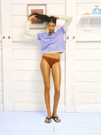 Model wearing striped polo shirt and brown briefs standing against white wood paneling for Guest In Residence