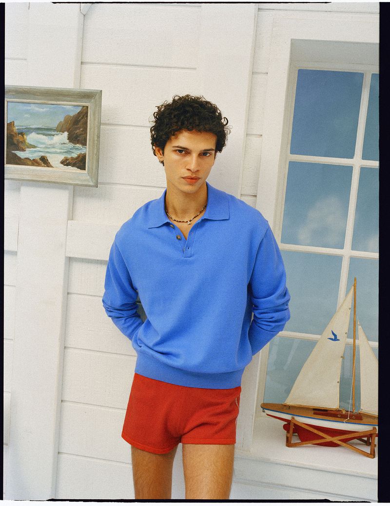 A male model wearing a blue polo sweater and red shorts stands indoors against white wood paneling.