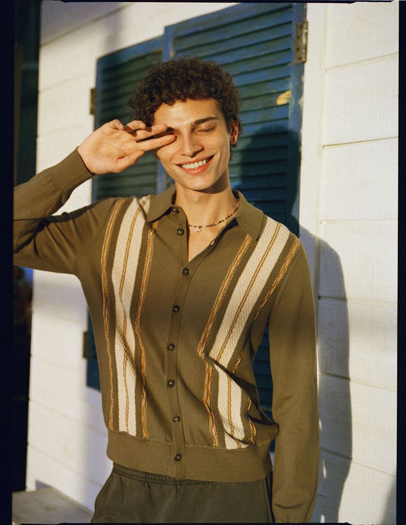 Bradley Veragten smiling with his hand over one eye, wearing an olive striped knit polo cardigan against a white wall with blue shutters.