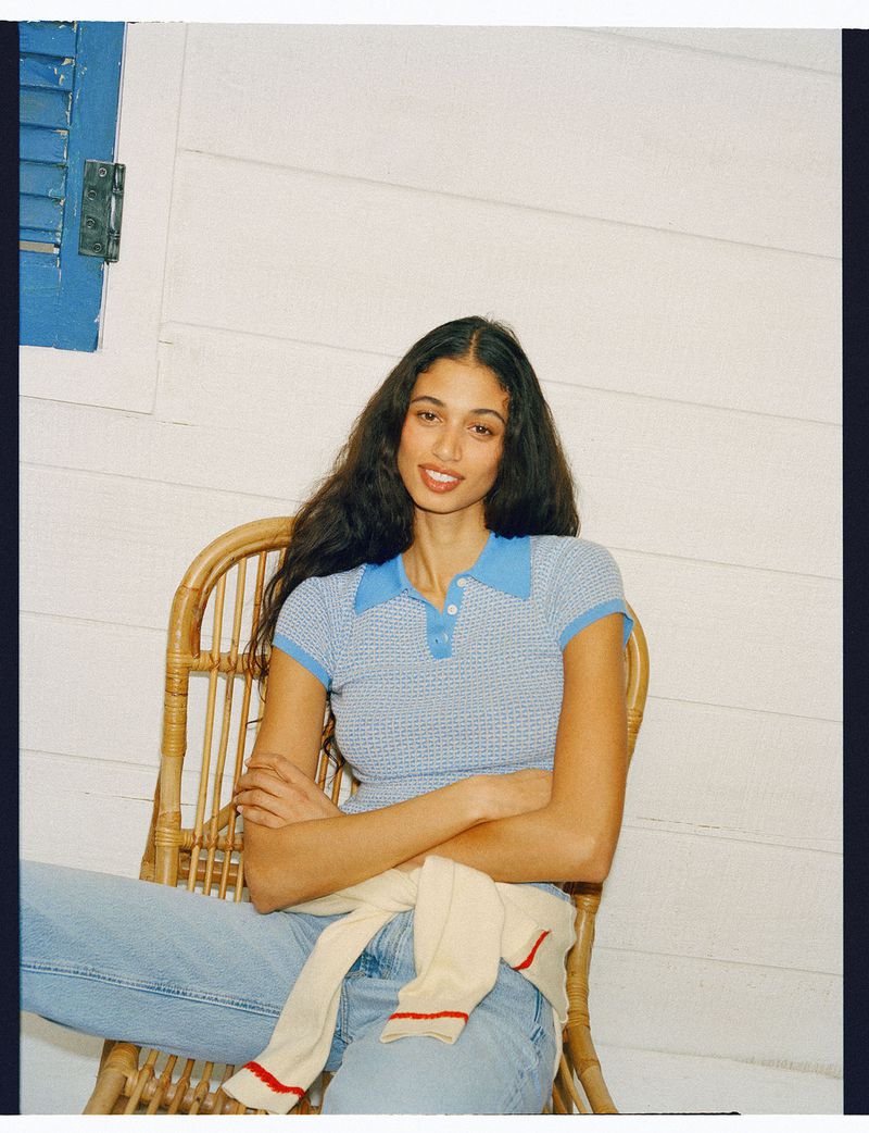 Model wearing a blue patterned polo and light wash jeans seated in a wicker chair against a white paneled wall