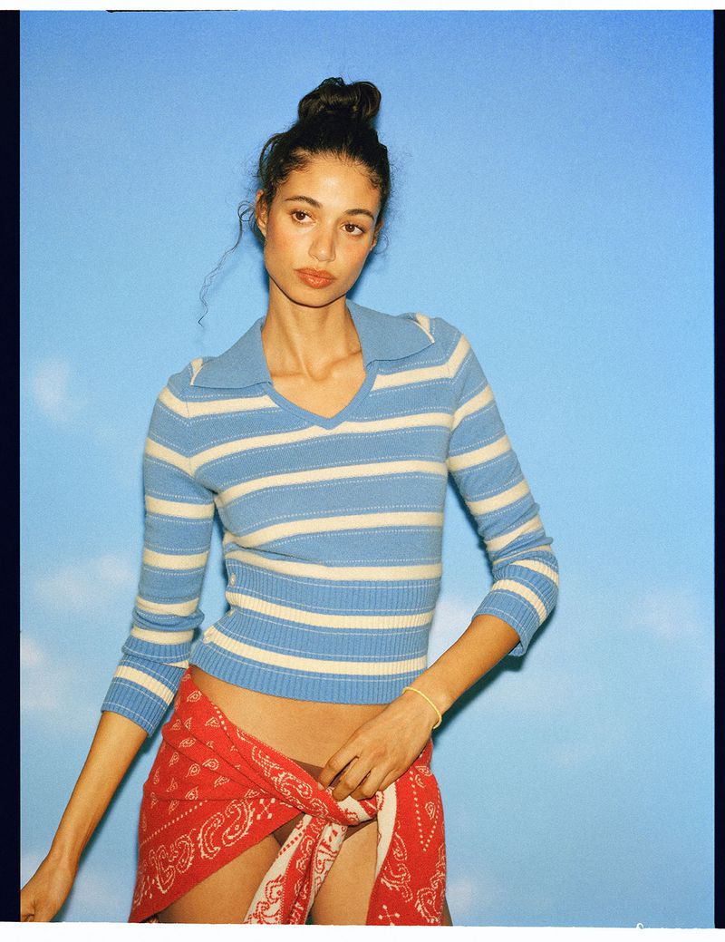Model Malika El Maslouhi posing in a blue and white striped knit polo top and red bandana wrap against a blue sky backdrop.