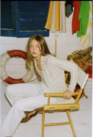 Gigi Hadid seated on a director chair wearing striped button-down shirt and white trousers against a coastal background