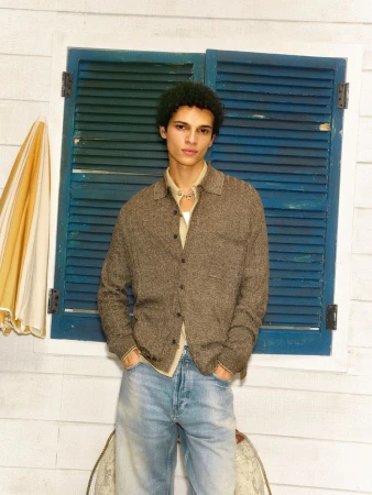 Model Bradley Veragten wearing a brown knit button-up shirt and light-wash jeans in front of blue wooden window shutters.