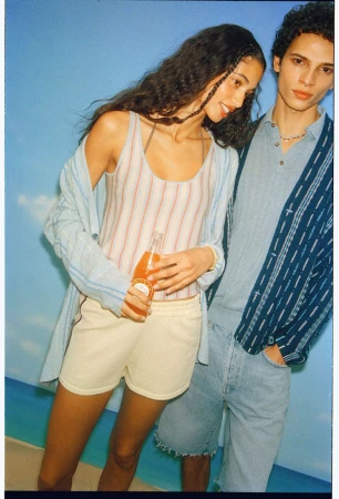 Model Malika El Maslouhi in a striped knit top holding a soda bottle next to Bradley Veragten against a painted sky backdrop.