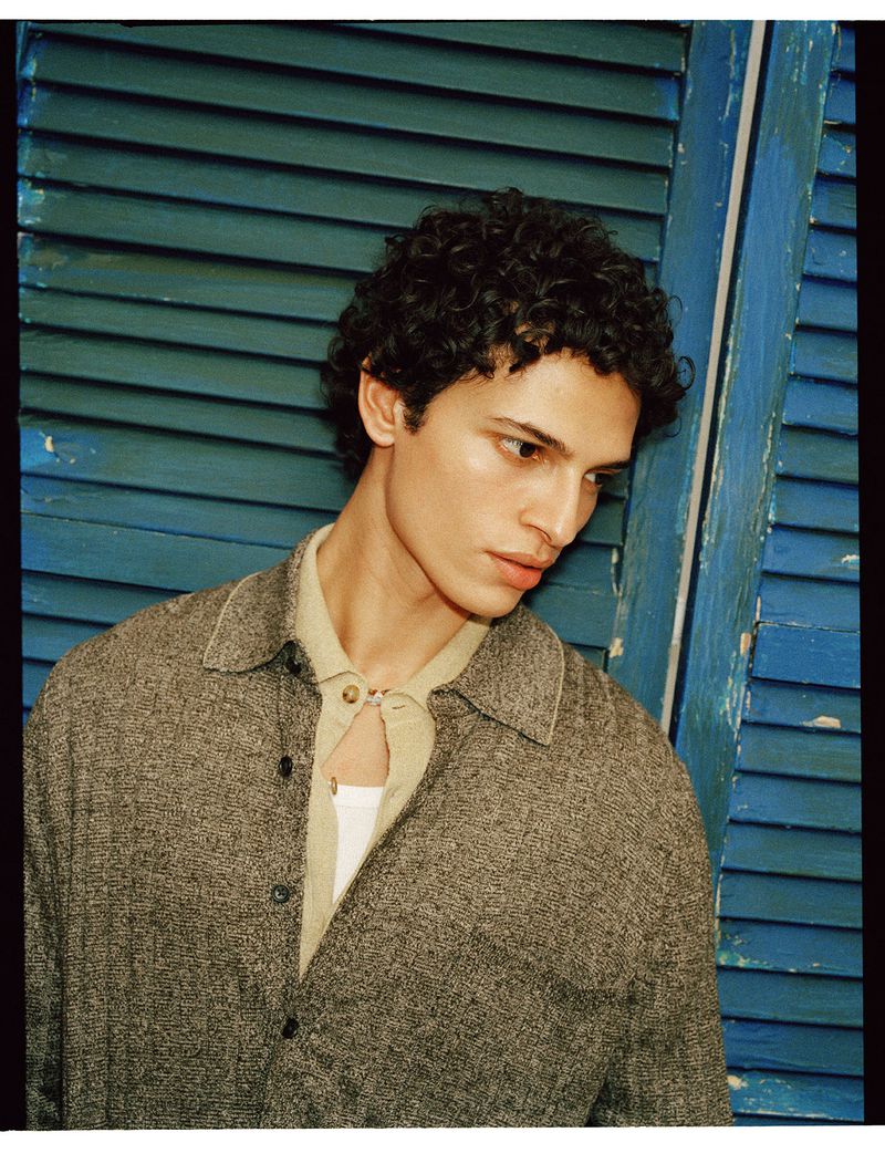 A young adult male poses in a grey button-down shirt and light-colored collared shirt in an indoor shot with a blue shutter background.
