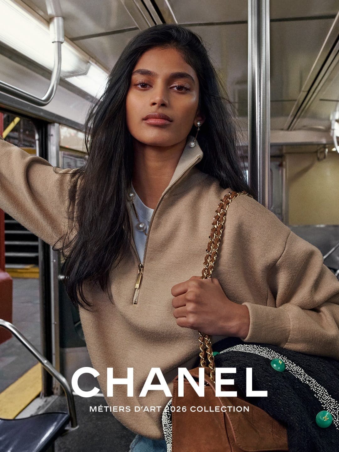 Model Bhavitha Mandava wearing a beige quarter-zip sweater and holding a Chanel chain handbag inside a subway car.