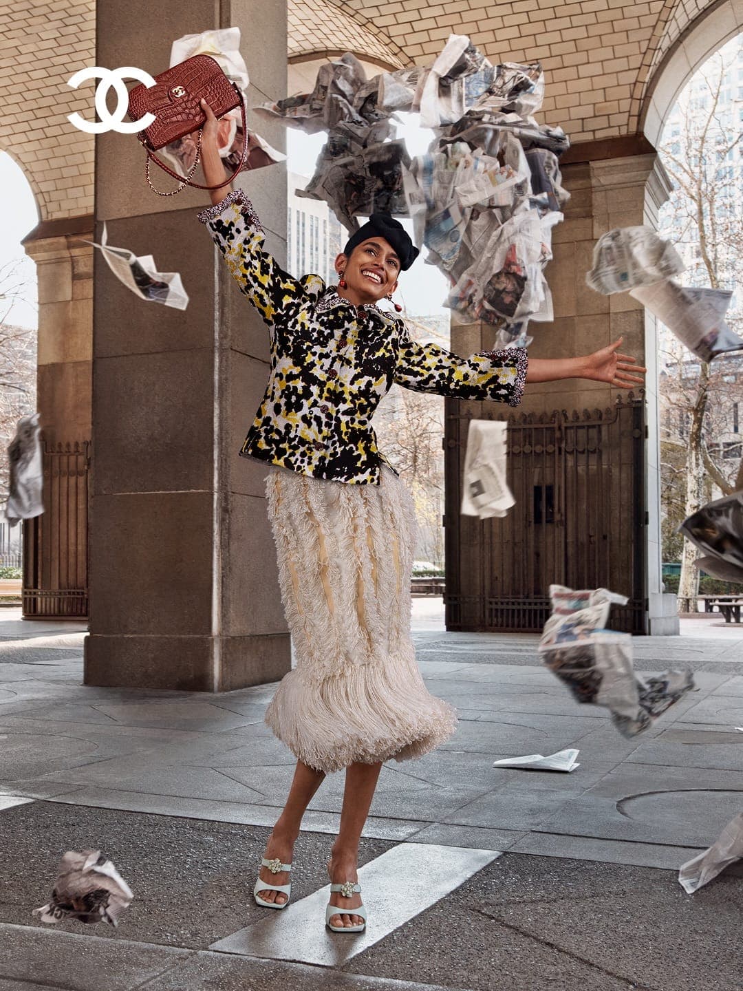 Smiling woman in a printed jacket and white fringe skirt holds a quilted handbag as newspaper pages fly through an arched colonnade.