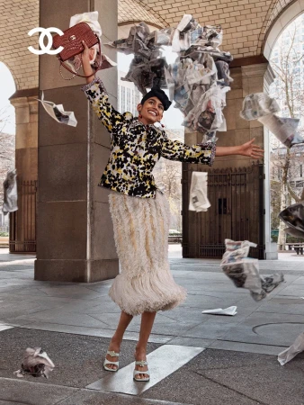 Smiling woman in a printed jacket and white fringe skirt holds a quilted handbag as newspaper pages fly through an arched colonnade.