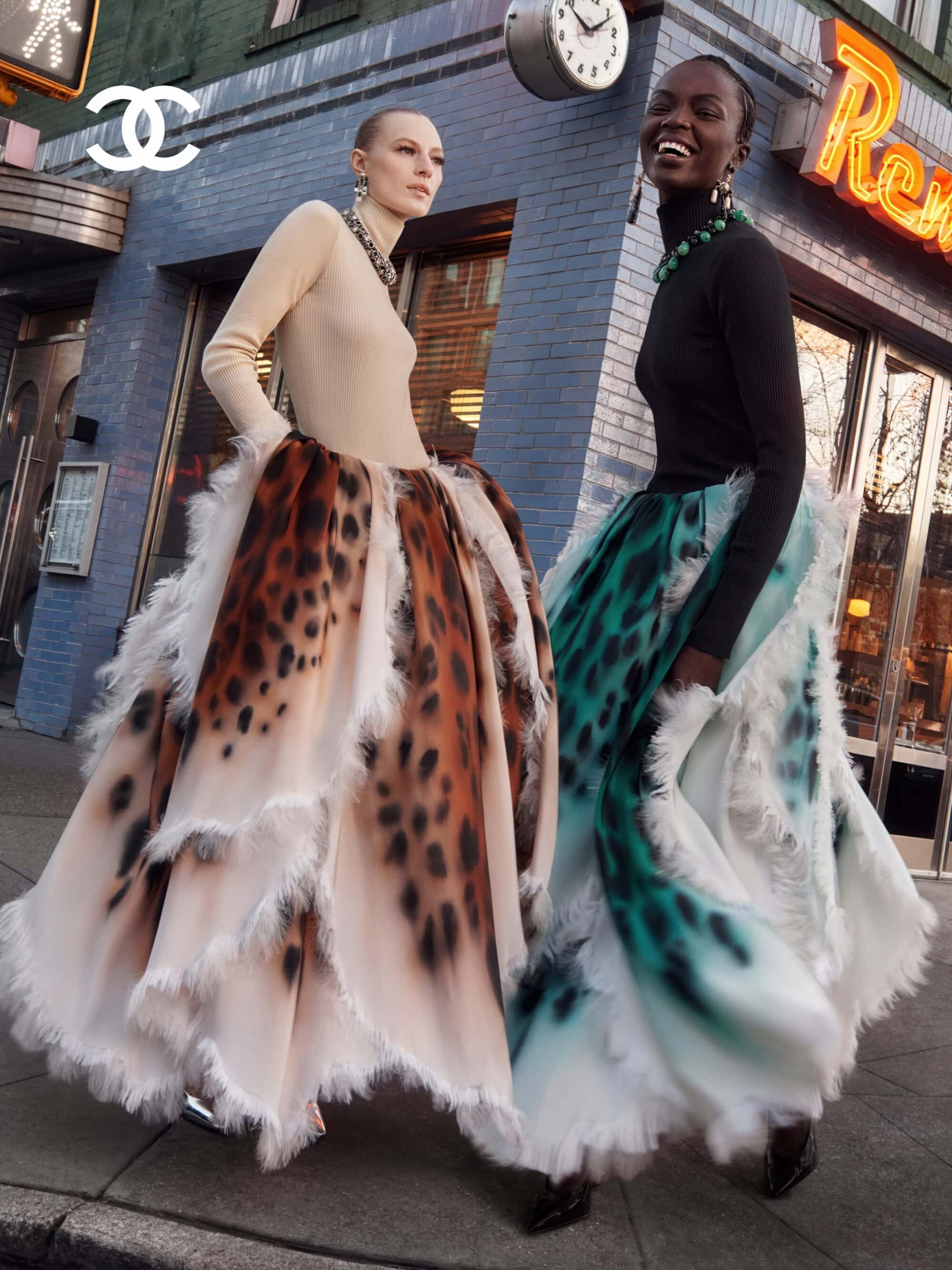 Two fashion models in high-neck tops and voluminous printed skirts posing on an urban sidewalk corner in front of a brick building.