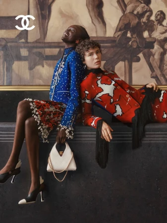 Two female models posing back to back against a dark wall with a large painting, wearing patterned jackets and holding a white handbag.