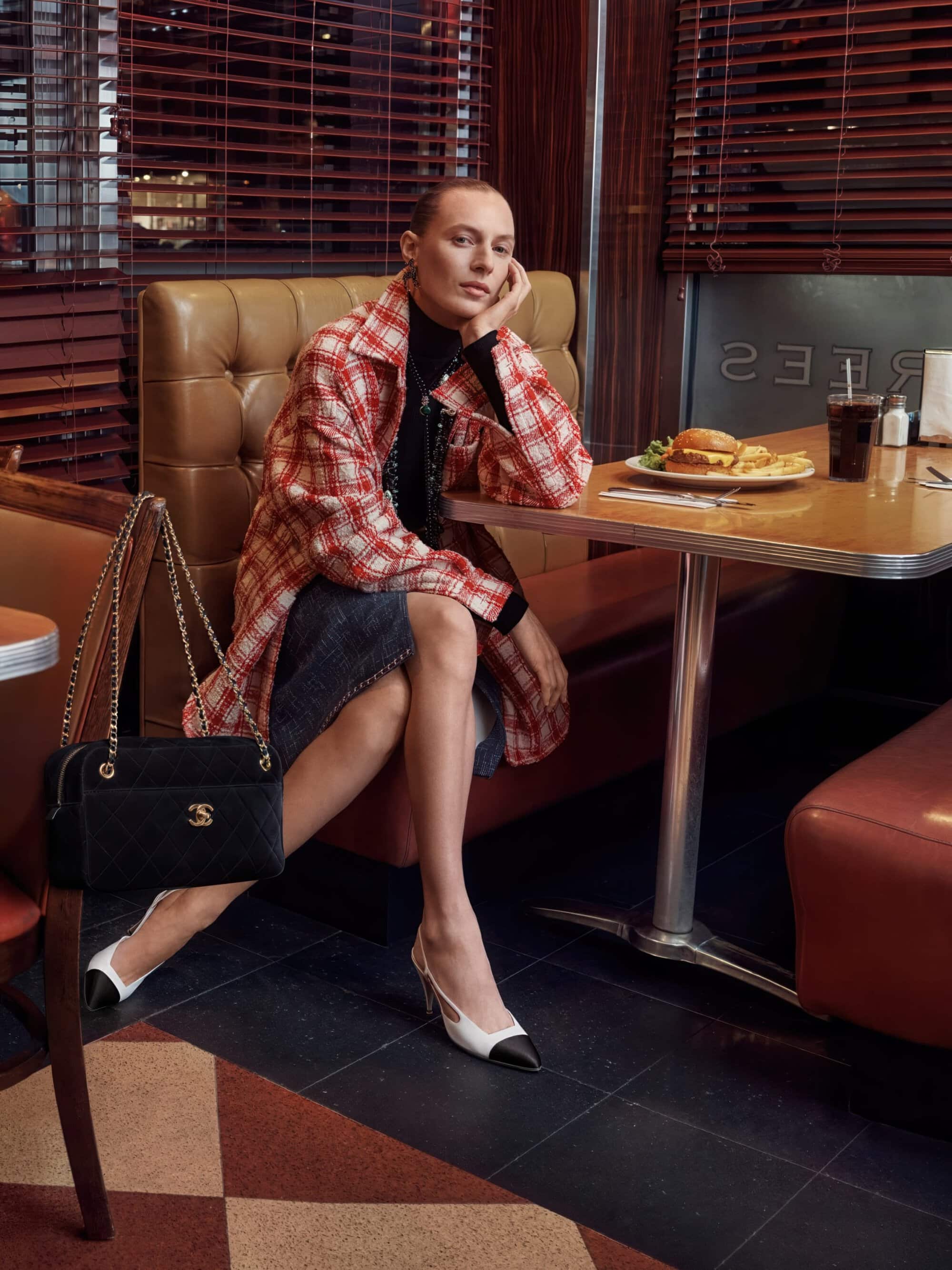 Julia Nobis seated in a diner booth wearing a red plaid tweed coat and two-tone slingback heels beside a black quilted handbag.