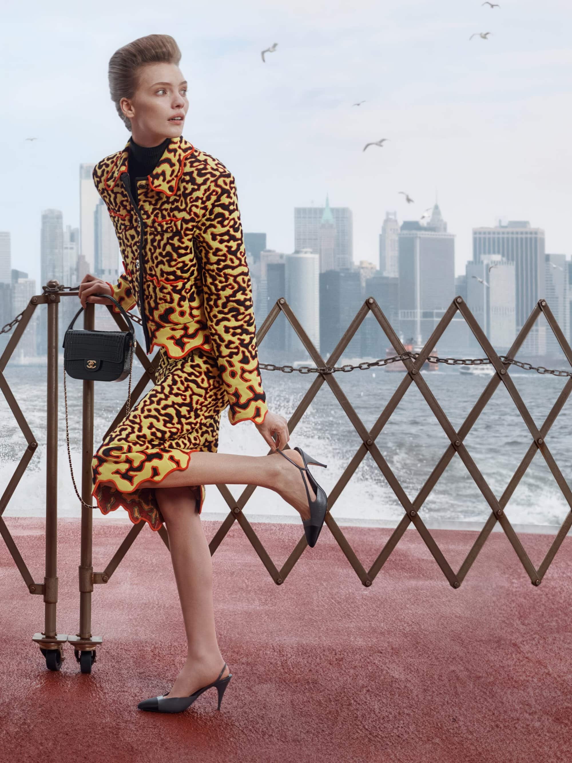 Model in a yellow and orange patterned tweed skirt suit adjusting her heel by a railing with a city skyline in the background.