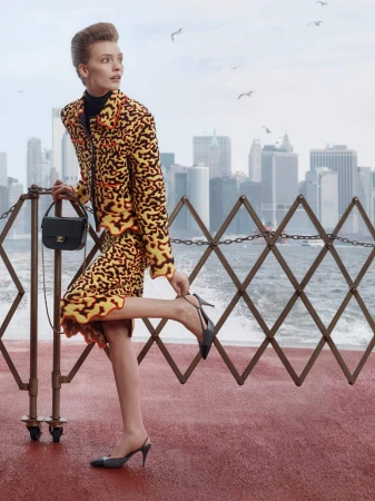 Model in a yellow and orange patterned tweed skirt suit adjusting her heel by a railing with a city skyline in the background.