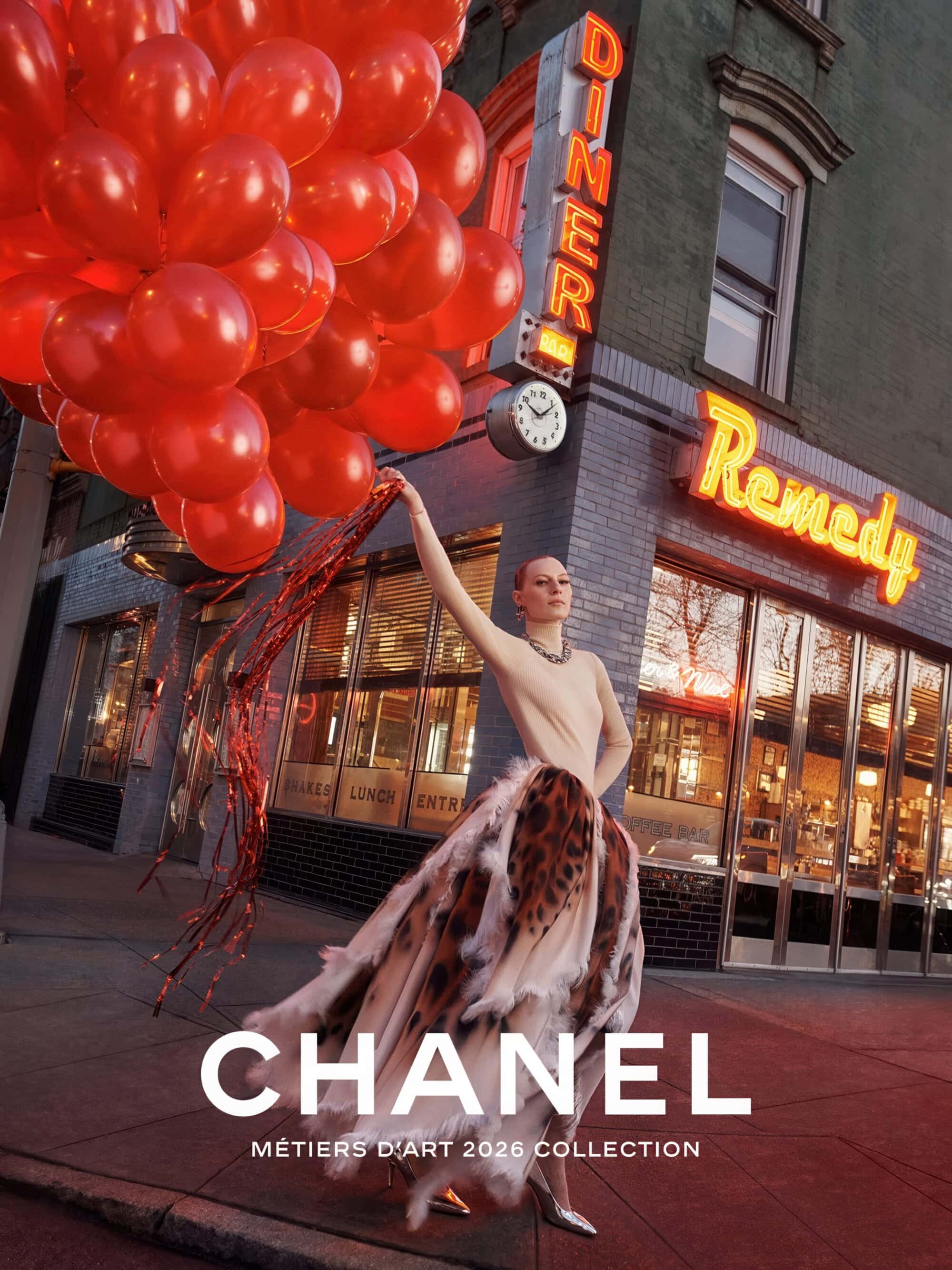 Fashion model holding a large bunch of red balloons on a city sidewalk in front of a neon diner sign for Chanel Métiers d'Art 2026.