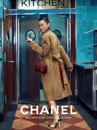 Model in quilted beige coat carrying a burgundy handbag in front of a blue tiled diner kitchen door
