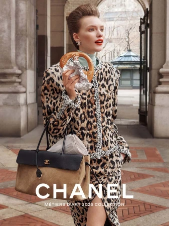 Model wearing a leopard print tweed suit and holding a pretzel while carrying a two-tone Chanel handbag in a stone courtyard.