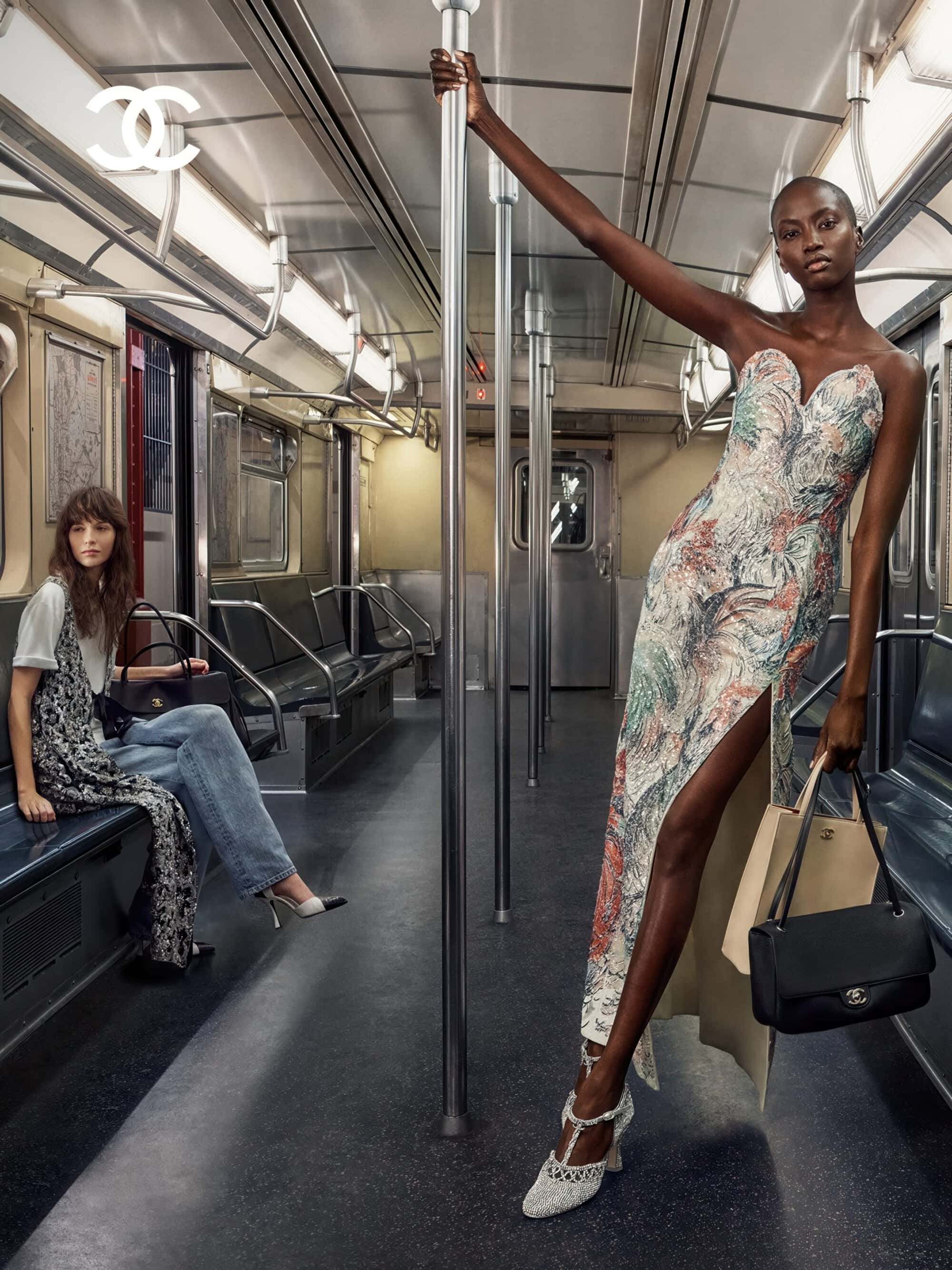 Model Anok Yai in a strapless patterned gown poses holding a subway pole next to a seated woman in a Chanel ad campaign.