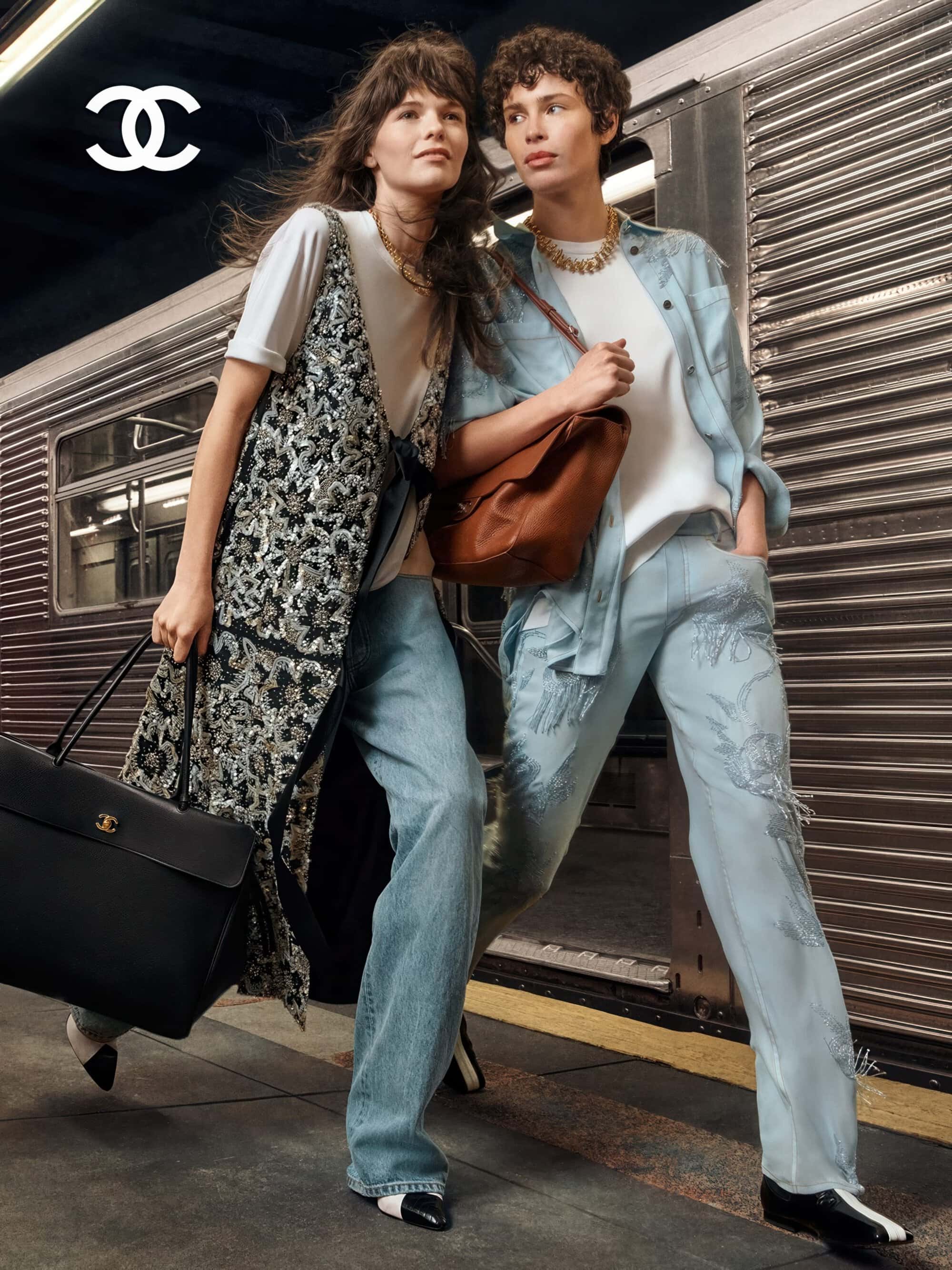 Two fashion models walking past a subway train wearing embroidered denim, white shirts, and carrying leather Chanel bags.