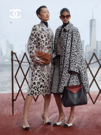 Two fashion models posing in patterned coats with Chanel leather handbags against a misty waterfront city skyline.