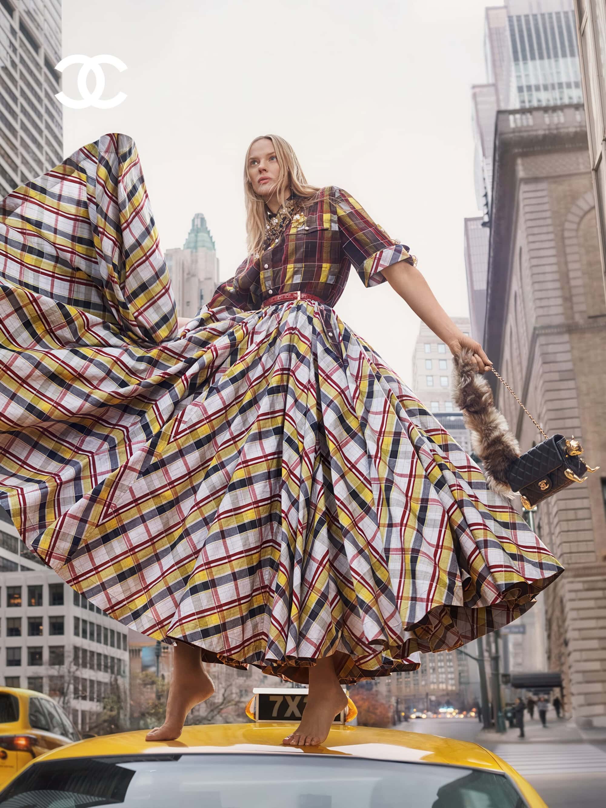 Fashion editorial featuring a female model in a plaid dress standing on a yellow taxi in a cityscape setting, accessorized with a handbag.
