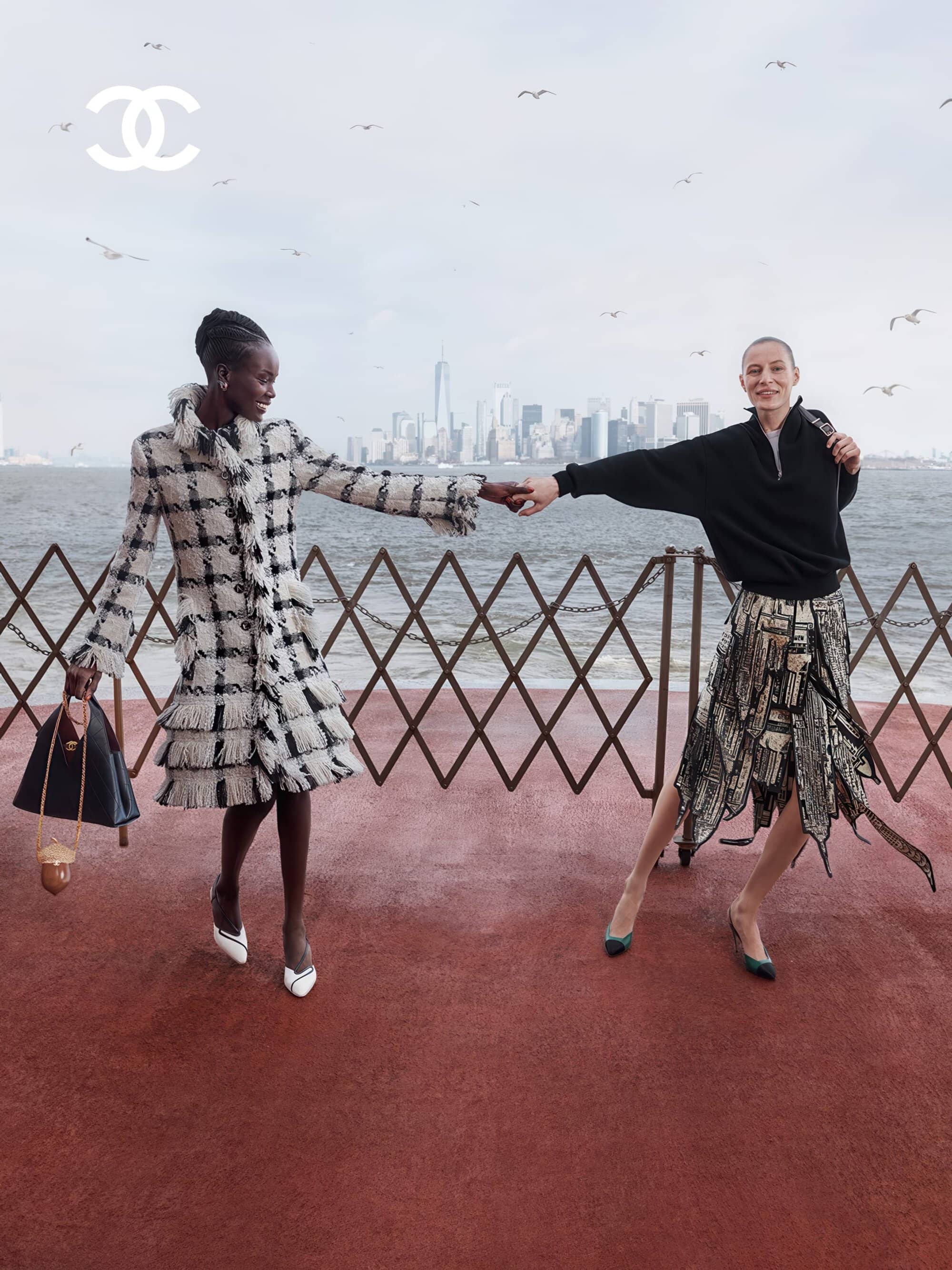 Anok Yai and Julia Nobis holding hands on a waterfront deck overlooking a city skyline in Chanel Métiers d'Art campaign.