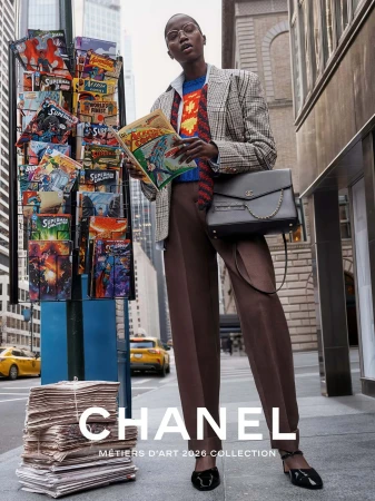 Model standing on a city sidewalk holding a vintage comic book next to a newsstand and a stack of newspapers.