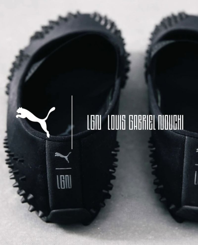 Close-up of black spiked Puma x LGN Louis Gabriel Nouchi Mostro slip-on shoes placed on a smooth grey surface.