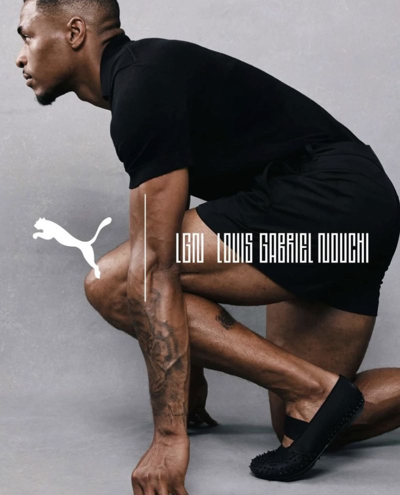 Muscular Black man in a black polo and shorts crouching in a sprinter pose against a grey studio backdrop.
