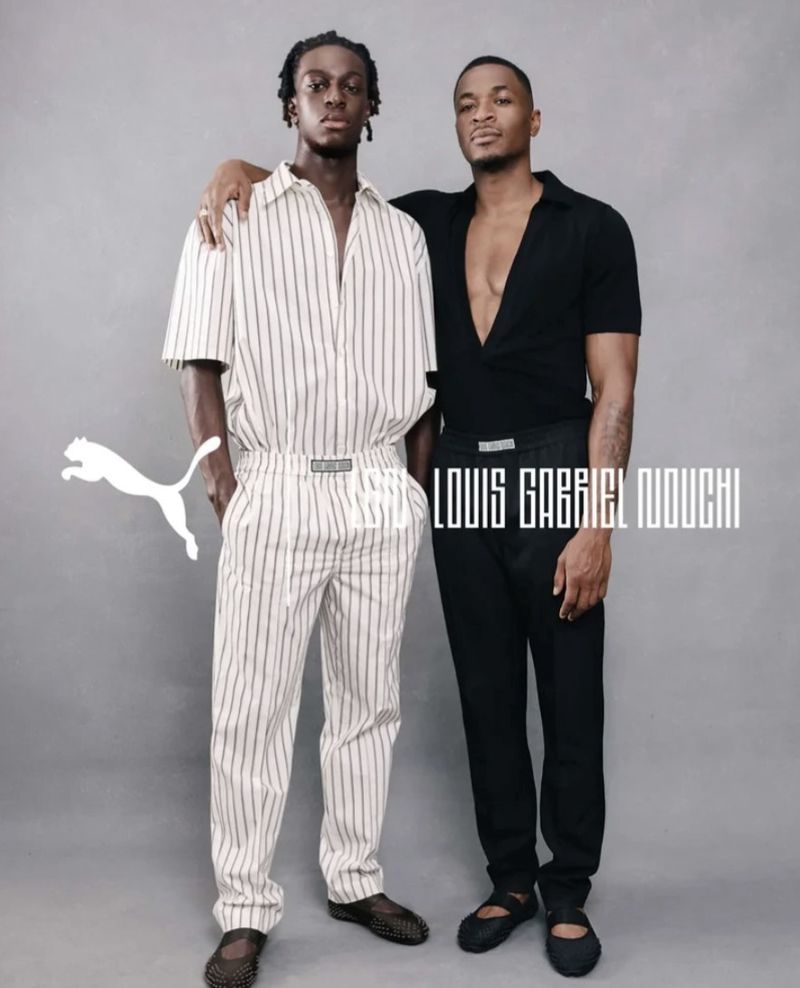 Two men posing together in studio wearing a striped matching set and an all-black outfit against a grey seamless backdrop.