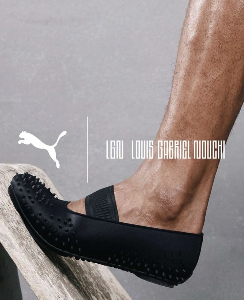 Close-up of a bare foot wearing a black spiked Puma x LGN Mostro flat shoe resting on a rustic wooden plank against a grey studio backdrop.