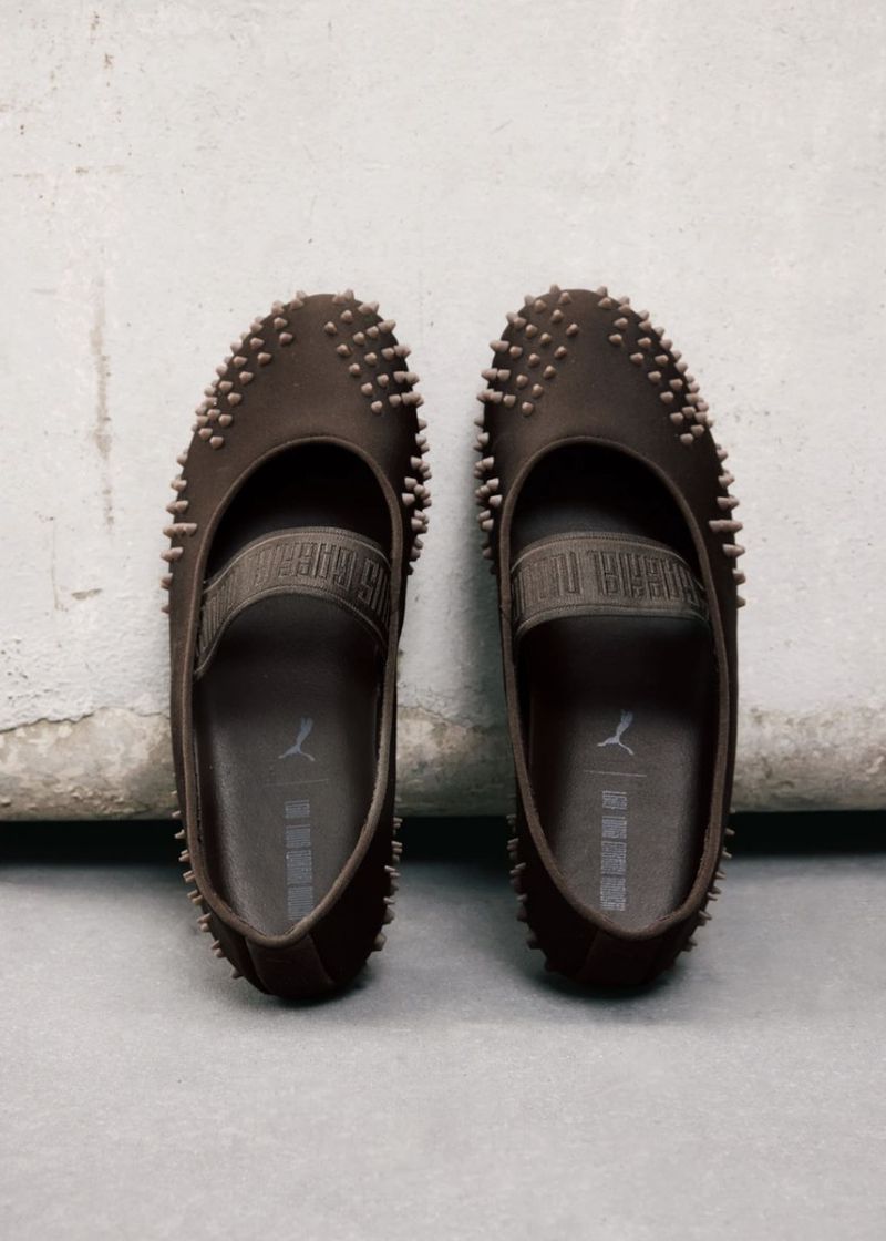 Pair of dark brown spiked ballet flat shoes from the Puma X LGN Mostro collaboration resting on a light grey concrete surface.